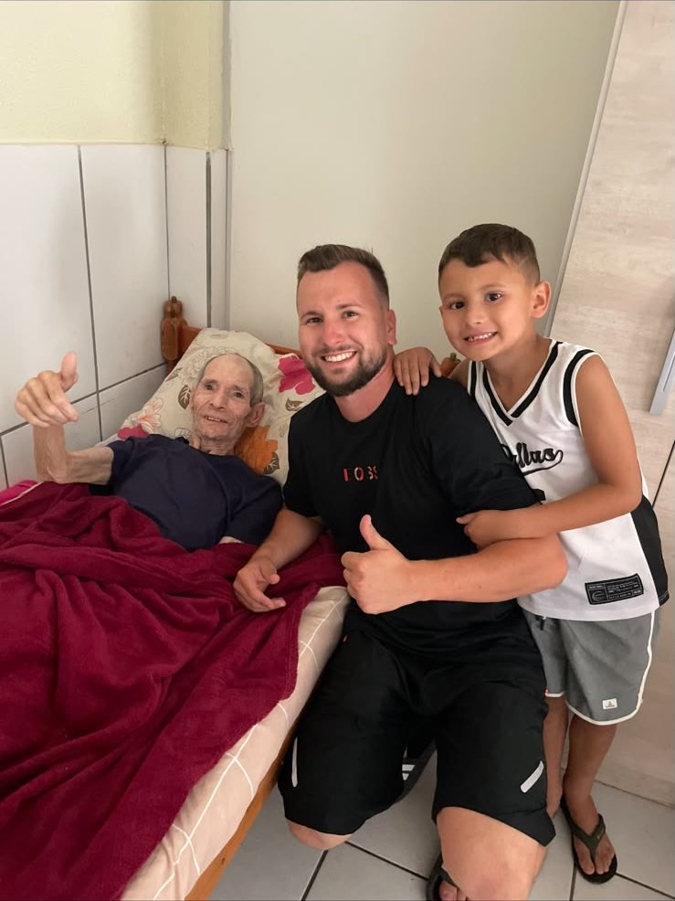 Morador acamado fazendo sinal de positivo ao lado de Renan e do filho